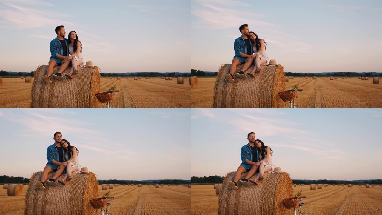 手持慢动作镜头的成年夫妇坐在田野里的干草捆上，拥抱并欣赏日落