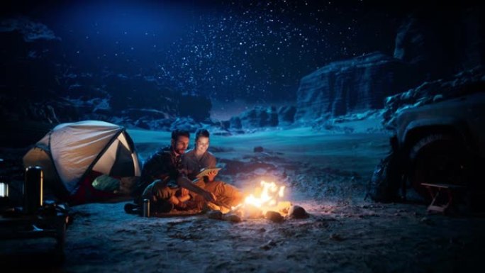 快乐的情侣晚上在峡谷露营，使用数字平板电脑，坐在篝火旁。两个旅行的人玩得开心，微笑，在社交媒体上发帖