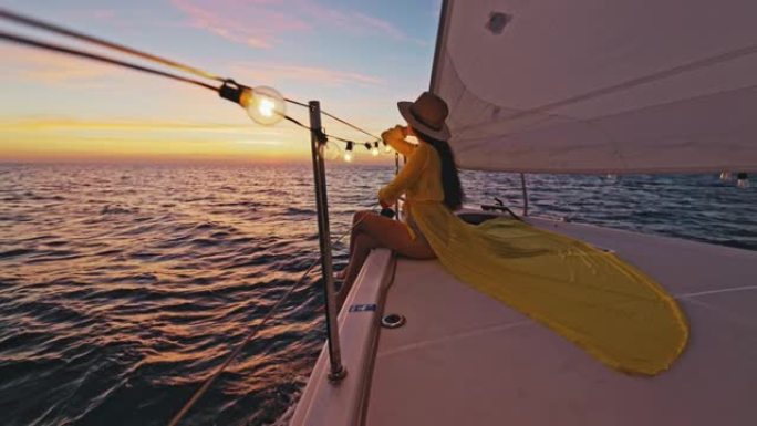 穿着黄色长裙的SLO MO女人坐在日落时航行的帆船甲板上