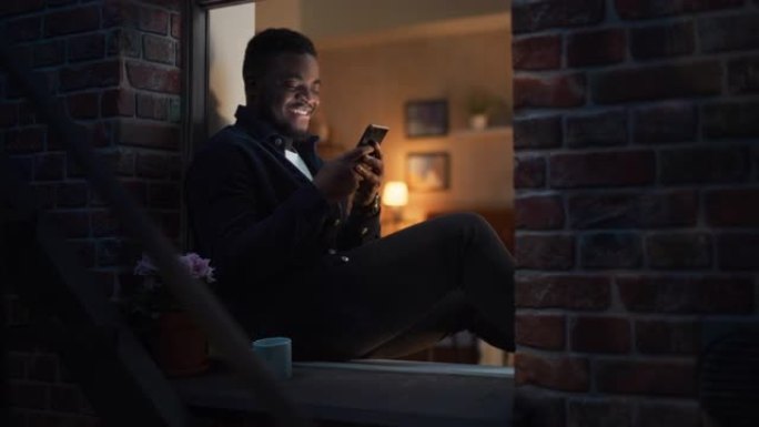 快乐的黑人在晚上坐在窗台上时，使用智能手机浏览有趣的在线内容或在社交媒体上与朋友聊天。男性笑着玩得开