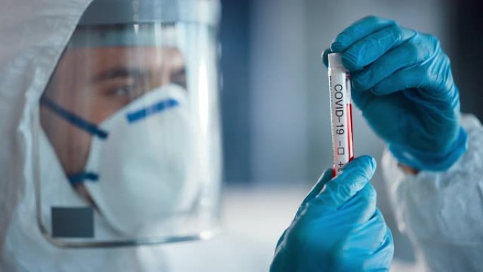 医学研究科学家穿着工作服，手术手套，面罩和盾牌将试管与血液样本和标签读取冠状病毒保持在一起。微生物实