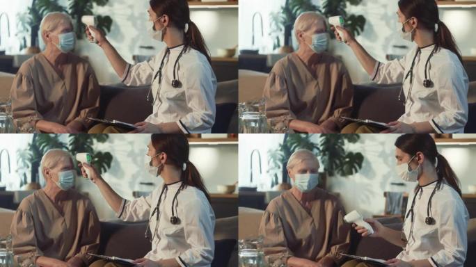 年轻的女医生戴着医用口罩，外套在家用红外体温计测量70岁快乐女性的体温。