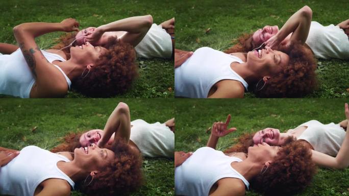 两名多民族妇女躺在公园草坪上，戴着耳机听音乐。黑人少女在大热天晒日光浴时与朋友分享她最喜欢的歌曲