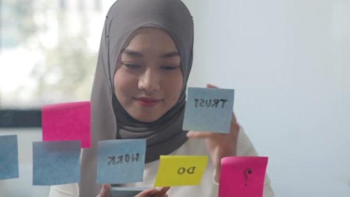 亚洲穆斯林女士写信息在新的普通办公室的玻璃板上贴一张便签。