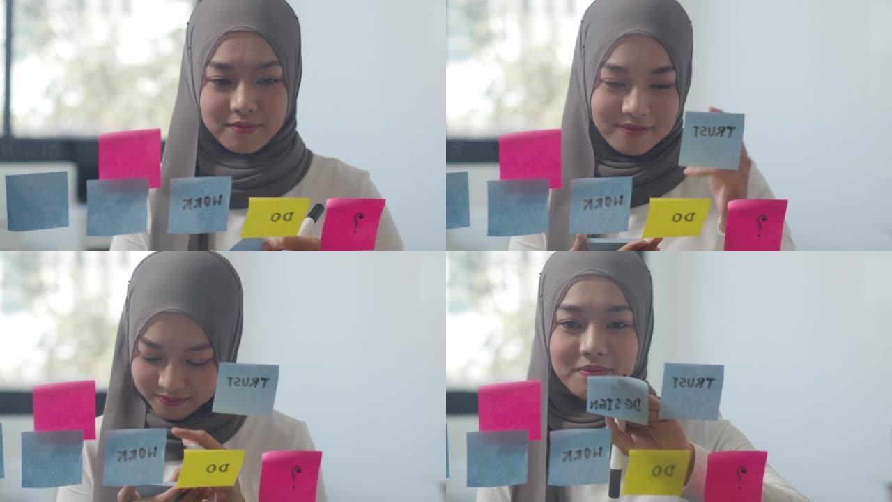 亚洲穆斯林女士写信息在新的普通办公室的玻璃板上贴一张便签。