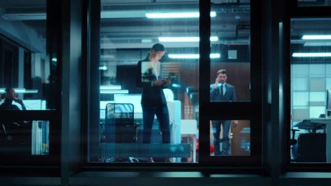 外面的镜头: 女商人在办公室的平板电脑上工作。经理站在窗边检查电子邮件。员工计划财务任务和业务发展。