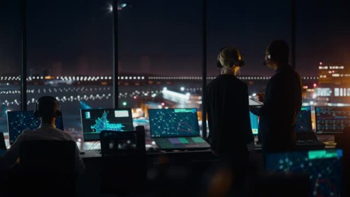 男女空中交通管制员带着耳机晚上在机场塔台交谈。办公室里满是台式电脑显示屏，配有导航屏幕，飞机飞行雷达