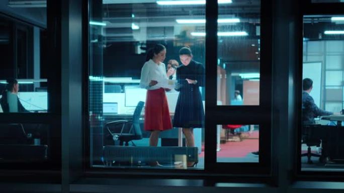 外面的镜头: 两名女商人在办公室的平板电脑上工作。经理站在窗边。员工谈论财务和业务发展。同事在他们身