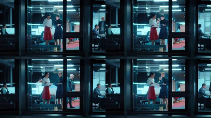 外面的镜头: 两名女商人在办公室的平板电脑上工作。经理站在窗边。员工谈论财务和业务发展。同事在他们身