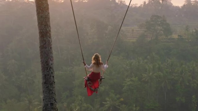 旅行女人在日出时在热带雨林上摇摆女游客坐在秋千上欣赏风景享受假期自由享受快乐假期生活方式慢动作