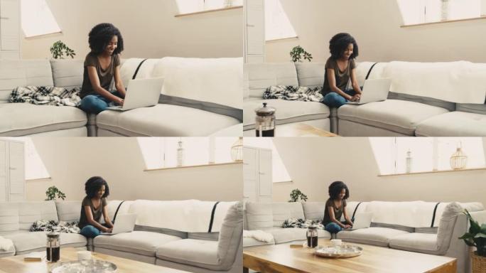 一名年轻的非洲裔美国妇女坐在家里的沙发上使用笔记本电脑。一位美丽的混血女性，非洲裔在客厅的沙发上放松