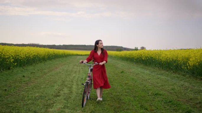 慢动作，追踪一个穿着红色连衣裙的黑发女郎的镜头，她骑着自行车穿过一片充满黄色金鱼草的草地