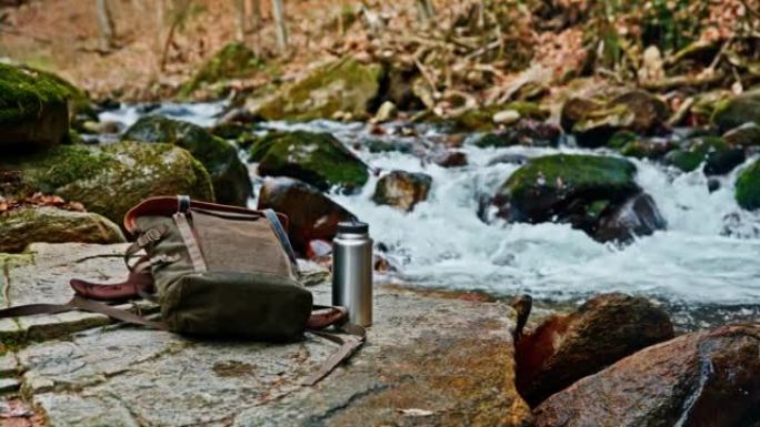 保温瓶与背包在岩石旁边流过森林小溪