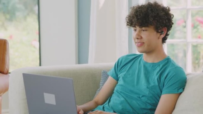 十几岁的男孩戴着无线耳塞坐在家里的沙发上，用慢动作拍摄的笔记本电脑进行视频通话