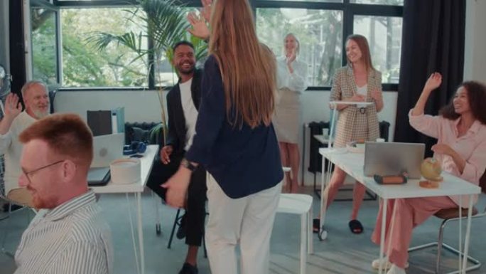 摄像机跟随快乐兴奋的女商人带着成功庆祝舞蹈进入办公室，团队鼓掌慢动作。