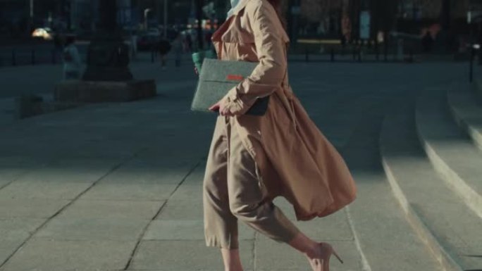 穿着优雅正装的成功美丽年轻女性首席执行官带着笔记本电脑慢动作微笑着走下城市台阶。