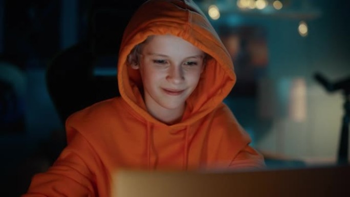 穿着连帽衫的可爱的小男孩在家里舒适的黑暗房间里使用笔记本电脑。快乐少年在线浏览教育研究，与朋友聊天，