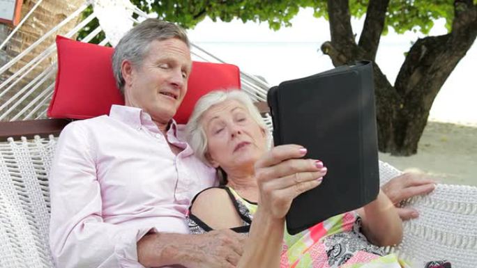 高级夫妇使用数字平板电脑在海滩吊床上放松