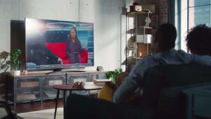 黑人夫妇在家里的沙发上看电视早间新闻时互相交谈。恋爱中的人共度时光。后视图移动镜头