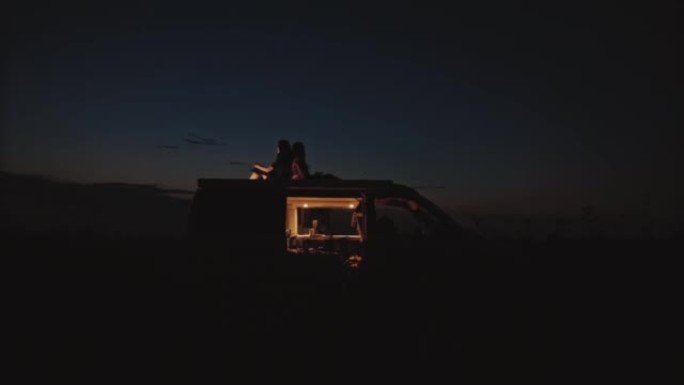 DS两名妇女晚上在田野中央背靠背坐在露营者的屋顶上