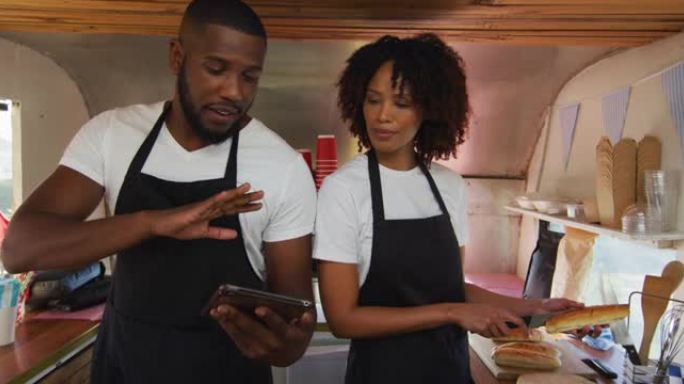非裔美国夫妇穿着使用数字平板电脑的围裙，并在餐车中准备热狗