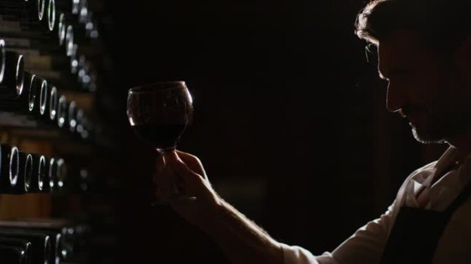 成功的男性酿酒师或侍酒师的剪影正在品尝酒窖中倒入透明玻璃杯中的红酒的味道并检查其质量。