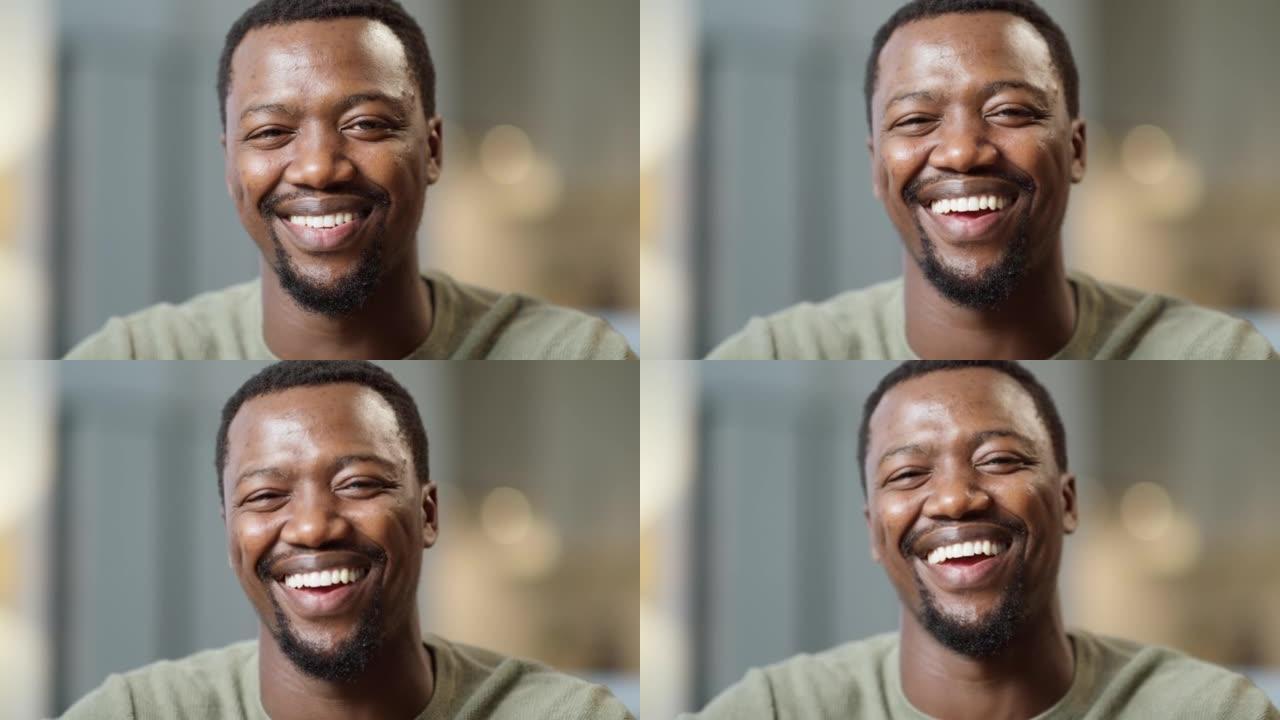 一个快乐的男人用惊人的牙齿微笑的肖像。英俊的非洲裔美国人坐在家里笑。快乐的黑人在家里放松，提供良好的