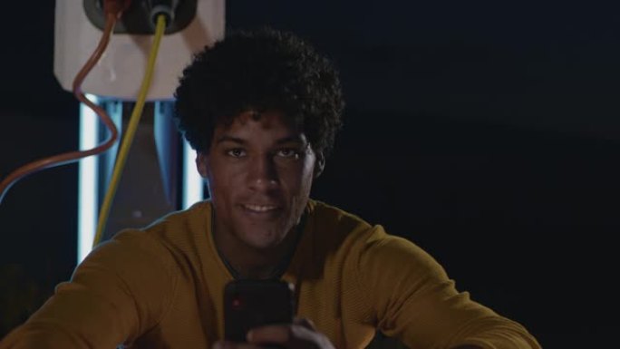 SLO MO肖像，一个年轻人在晚上在停车场上使用智能手机充电时使用智能手机