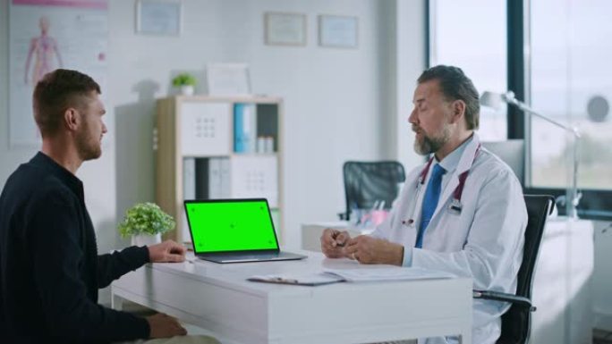 家庭医生正在健康诊所的绿屏计算机上向年轻患者解释诊断。穿着白大褂的助理正在医院办公室阅读病史。