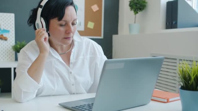 快乐的女商人在办公室里用笔记本电脑工作，通过耳机享受音乐