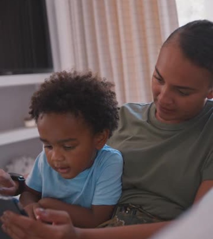 军队母亲穿着制服坐在沙发上，儿子在玩游戏或在平板电脑上播放电影——用慢动作拍摄
