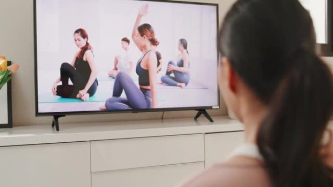 穿着运动服的年轻亚洲女性在地板虚拟健身课上的垫子上与一群人在电视上的视频会议上练习瑜伽，在家里的客厅