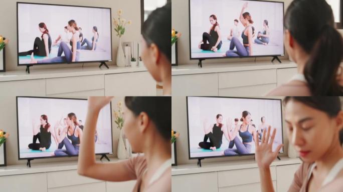 穿着运动服的年轻亚洲女性在地板虚拟健身课上的垫子上与一群人在电视上的视频会议上练习瑜伽，在家里的客厅