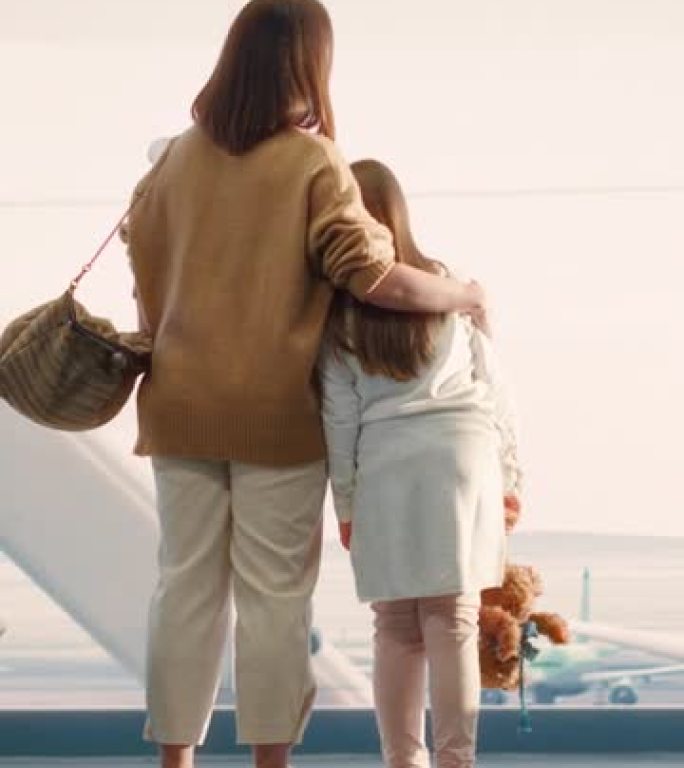 垂直屏幕。机场航站楼: 美丽的母亲和可爱的小女儿等待度假航班，看着窗外的飞机。航空枢纽登机休息室的年