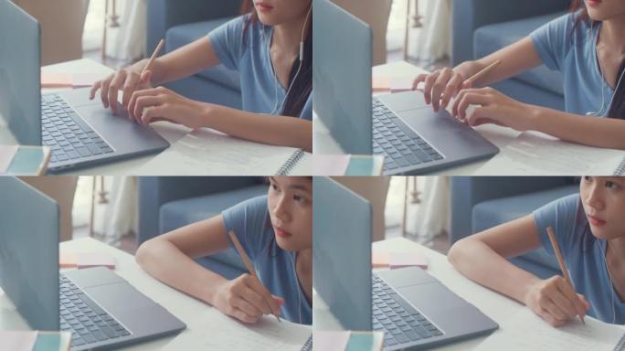 年轻的亚洲女孩少年，戴着休闲耳机，使用笔记本电脑在线学习在家里的客厅写讲座笔记本。隔离教育在线电子学