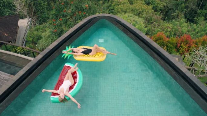 两个女人漂浮在游泳池里躺在充气玩具上最好的朋友暑假在热带酒店度假时玩得开心，穿着比基尼顶视图