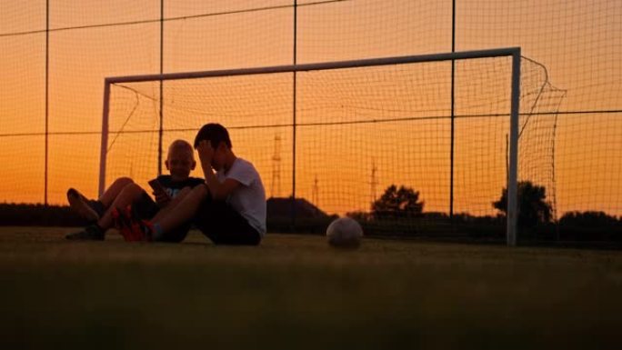 男孩在黄昏时坐在草地上与球一起在草地上使用手机