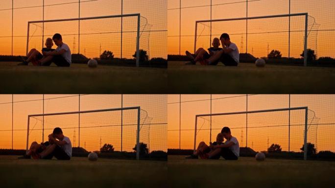 男孩在黄昏时坐在草地上与球一起在草地上使用手机