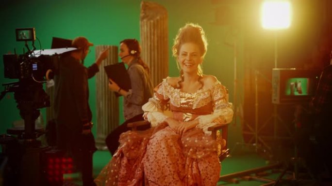 在古装戏剧电影中: 美丽的微笑女演员穿着文艺复兴时期的连衣裙，快乐地坐在椅子上，背景是绿色的屏幕。工