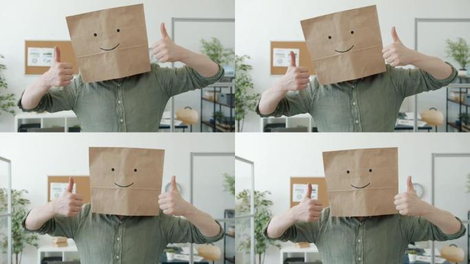 商人的肖像穿着带有微笑表情符号的面包袋，在工作场所表现出竖起大拇指的手势