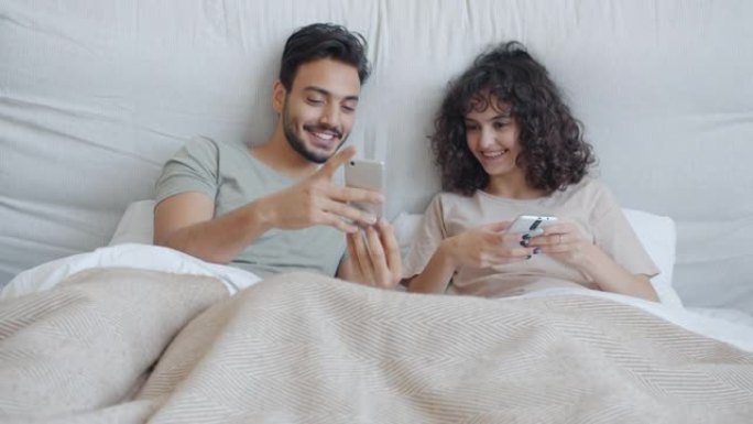 快乐的混血夫妇使用智能手机在家里的床上聊天和大笑放松