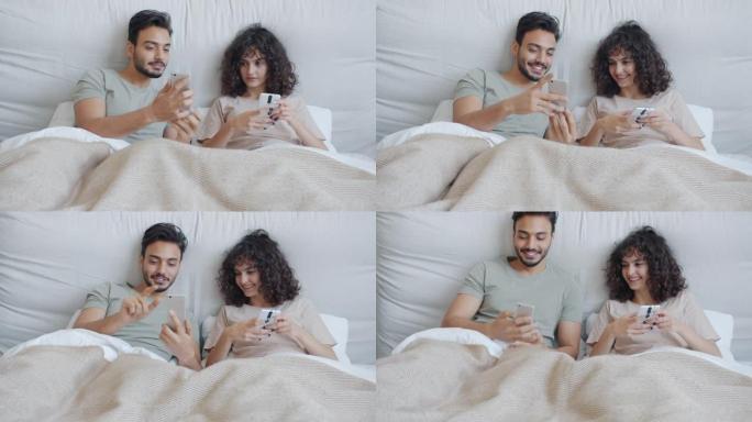 快乐的混血夫妇使用智能手机在家里的床上聊天和大笑放松