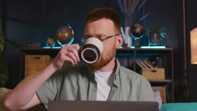 在家工作年轻快乐微笑的白人商人戴眼镜，在舒适的客厅用笔记本电脑啜饮咖啡。