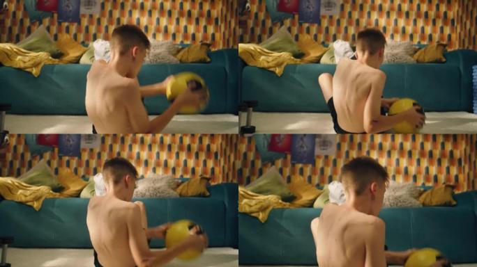 运动男孩做上半身锻炼与足球坐在他的房间的地板上