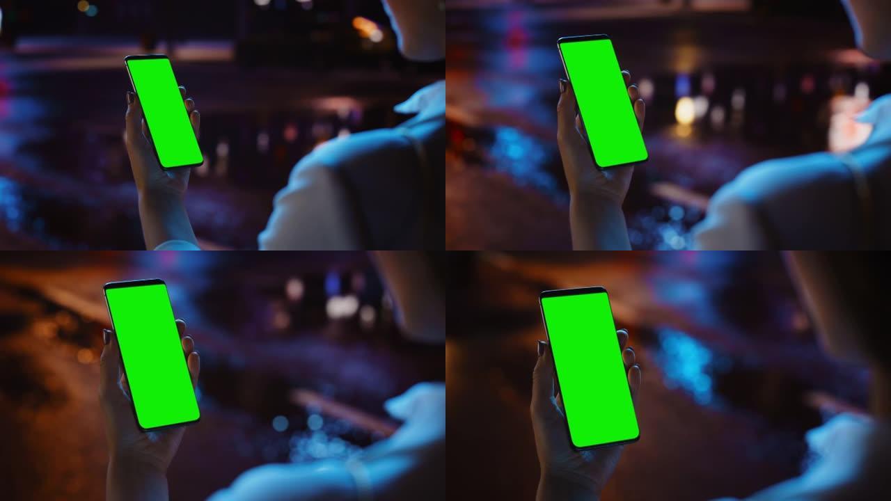 女人站着时使用色度键智能手机，看着绿色屏幕。充满霓虹灯的夜城街道。女性使用手机。肩膀上的特写镜头追踪