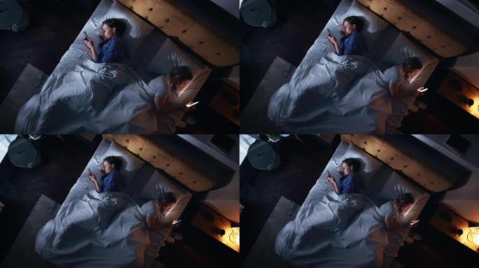 顶视图公寓卧室: 白人年轻夫妇躺在床上，使用智能手机。两口之家在睡觉前使用手机浏览社交媒体，搜索互联