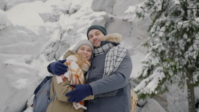 站在冬季森林里的快乐年轻夫妇的慢动作肖像，微笑着看着相机
