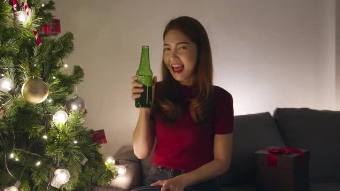 年轻的亚洲女性喝啤酒玩得开心，快乐的夜晚派对视频通话与情侣交谈，家里客厅装饰有装饰品的圣诞树。