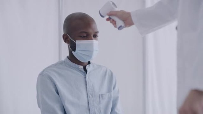 一名男性医生在非洲人的公寓中使用非接触式红外温度计测量患者的体温，该男子戴着口罩。医生在做新型冠状病