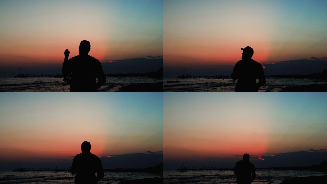 快乐旅游男人的史诗般的电影剪影，带着相机在海边享受令人惊叹的红霞，向前走。